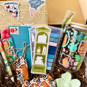 May include: A gift box filled with stationery and accessories. Includes a pen, keychain, coasters, stickers, and a jar with a straw. The items feature a capybara and floral design, with a green and blue color scheme.