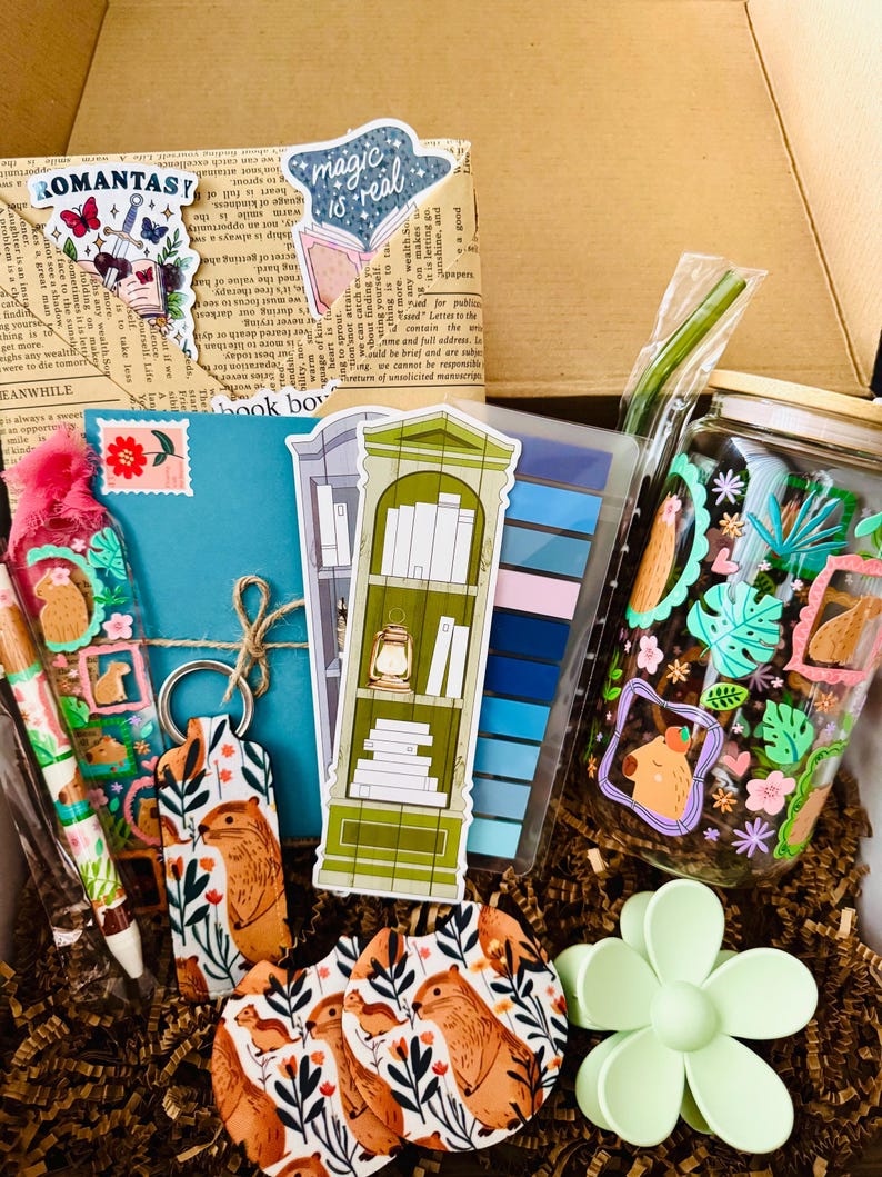 May include: A gift box filled with stationery and accessories. Includes a pen, keychain, coasters, stickers, and a jar with a straw. The items feature a capybara and floral design, with a green and blue color scheme.