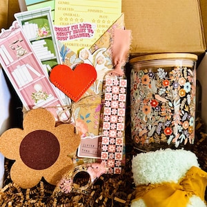 May include: A gift box containing a book review card, a floral glass tumbler with a bamboo lid, a flower-shaped cork coaster, a fuzzy light green cloth with a mustard ribbon, a pink bookmark, a red heart cutout, and various paper stationery items.