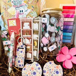May include: A gift box filled with stationery and decorative items. Includes a glass jar with a wooden lid, stickers, a pen, a keychain, coasters, and a pink flower-shaped clip. The items feature floral and vintage-themed designs.