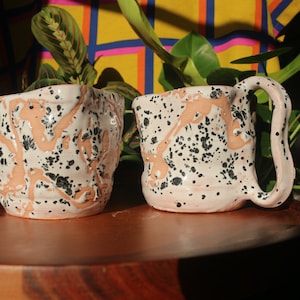 Puede incluir: Dos tazas de cerámica con base blanca y motas negras. Las tazas tienen una forma orgánica única y un esmalte moteado.