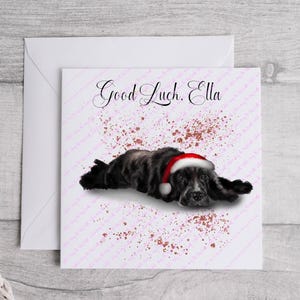 May include: A greeting card with a black and gray dog wearing a Santa hat, lying down. The card has the text "Good Luck, Ella" in cursive, with pink and red speckles. The card is inside a white envelope.