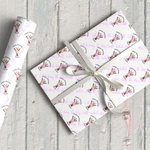 May include: A roll of white wrapping paper and a wrapped gift box, both featuring a repeating pattern of a giraffe with a pink floral crown and the text "Happy Birthday Queen". A white ribbon is tied around the box.