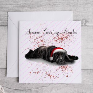 May include: A Christmas card featuring a black and gray dog wearing a Santa hat, lying down on a pink background with the words "Seasons Greetings Amelia". The card is inside a white envelope.