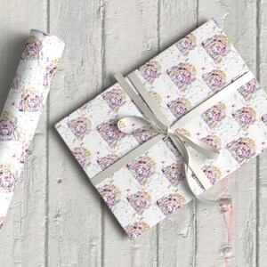 May include: White wrapping paper with a repeating pattern of watercolour illustrations of a dog's face. The dog has pink, purple, and yellow fur with white accents. The paper is rolled up and tied with a white ribbon.