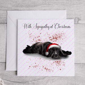May include: A greeting card with the text "With Sympathy at Christmas" features a black and gray dog wearing a Santa hat. The card has a pink and red speckled design and is placed on a white envelope.
