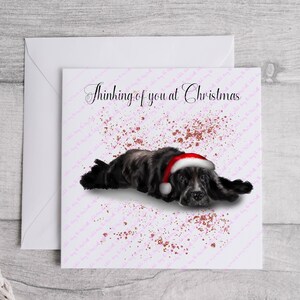 May include: A greeting card with a black and gray dog wearing a Santa hat, lying down. The card reads "Thinking of you at Christmas" in elegant script, with pink and gold accents.