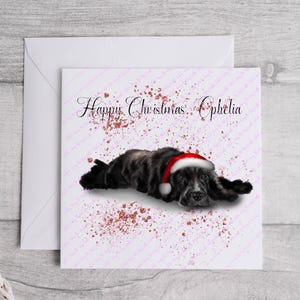 May include: A Christmas card featuring a black dog wearing a Santa hat, lying down on a white background with pink and gold accents. The card reads "Happy Christmas Ophelia" in elegant script.
