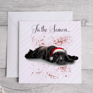 May include: A Christmas card featuring a black and gray dog wearing a red and white Santa hat. The card has the text "Tis the Season..." in elegant script, with a pink and brown speckled background.