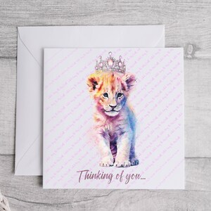 K&ouml;nnte beinhalten: Eine Aquarell-Illustration eines L&ouml;wenjungen mit einer glitzernden Krone. Das Junge sitzt auf einem wei&szlig;en Hintergrund mit dem Text "Thinking of you..." darunter.