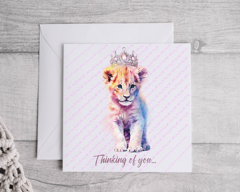 K&ouml;nnte beinhalten: Eine Aquarell-Illustration eines L&ouml;wenjungen mit einer glitzernden Krone. Das Junge sitzt auf einem wei&szlig;en Hintergrund mit dem Text "Thinking of you..." darunter.