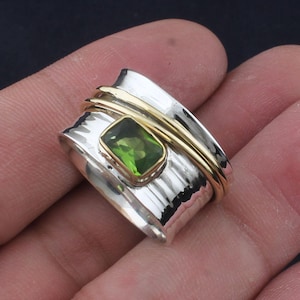 Könnte beinhalten: Ein drehbarer Silber- und Goldring mit einem grünen Edelstein. Der Ring ist texturiert und hat eine gehämmerte Oberfläche.