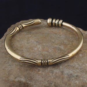 Puede incluir: Un brazalete de color dorado con diseños lineales y geométricos grabados. El brazalete abierto presenta bandas decorativas y una forma suave y redondeada. La joya se exhibe sobre una superficie de piedra texturizada de color marrón claro.