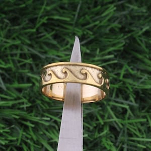 Może przedstawiać: Złoty pierścionek z wzorem fal. Pierścionek jest trzymany przez narzędzie.