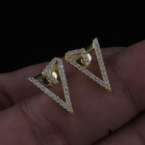 Puede incluir: Un par de pendientes dorados con forma triangular. Los bordes de los triángulos están bordeados con pequeñas piedras preciosas transparentes. Los pendientes se muestran sobre un fondo oscuro.