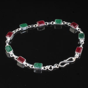 Peut inclure: Un bracelet en argent orné de pierres précieuses rectangulaires rouges et vertes alternées. Les pierres sont serties dans des chatons individuels en argent et reliées par de petits maillons en argent. Le bracelet a une fermeture à crochet. Le bijou est sur fond noir.