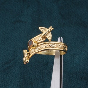 May include: Gold ring featuring a bee design and a brown gemstone. The ring is displayed against a blue backdrop.