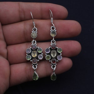 May include: A pair of silver dangle earrings featuring a cluster of colourful gemstones, including green, yellow, purple, and red. The earrings have a teardrop-shaped gemstone hanging from the bottom.