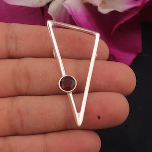 May include: A silver pendant in a triangular design, featuring a small, circular, deep red gemstone. The pendant is held against a skin background and a blurred floral arrangement.