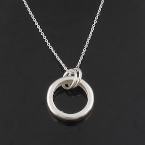 May include: A silver-colored necklace with a large, circular pendant. The pendant is connected to a delicate chain. The pendant has a smaller ring intertwined with the larger ring. The necklace is displayed against a black background.