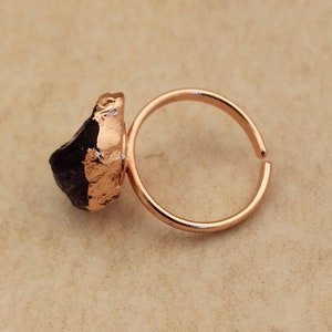 May include: A rose gold ring with a raw amethyst stone. The stone is a deep purple color and has a rough, natural texture. The ring is adjustable and can be worn on any finger.