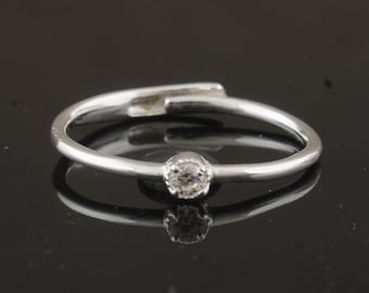 Op de afbeelding: Een zilveren ring met een enkele, ronde, heldere edelsteen. De band heeft een uniek, open ontwerp met een kleine opening aan de bovenkant. De ring staat tegen een donkere achtergrond, waardoor het reflecterende oppervlak wordt benadrukt.
