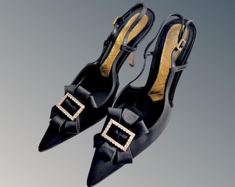 May include: A pair of black satin slingback heels with pointed toes. Each shoe features a decorative bow with a rectangular, rhinestone-studded buckle. The interior is gold with a quilted pattern, and the heels are approximately 2 inches high.