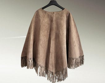 May include: A brown suede-textured poncho featuring a wide, loose-fitting silhouette with a rounded neckline and a vertical seam down the center. The hemline is finished with long, dangling fringe trim that hangs evenly around the entire bottom edge.