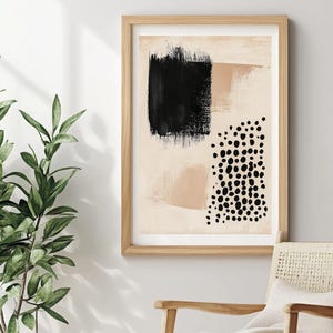 May include: Abstract art print in a wooden frame. The artwork features a black brushstroke, a tan shape, and a cluster of black dots on a beige background. The print is displayed near a woven chair and a potted plant.
