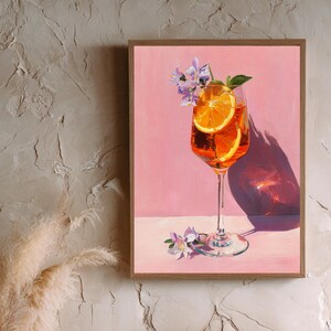 Op de afbeelding: Een ingelijst schilderij van een cocktail in een glas met sinaasappelschijfjes, paarse bloemen en een muntblad. De drank staat tegen een roze achtergrond met een wit oppervlak, in een houten lijst.