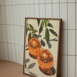 Puede incluir: Una impresi&oacute;n enmarcada de una pintura que representa dos bolas de discoteca naranjas colgando de una rama frondosa. El fondo es de color crema claro.