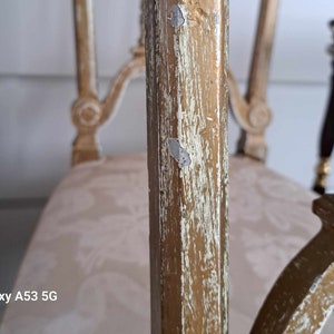 May include: Close-up of a gold-painted wooden chair with a white fabric seat. The paint is chipped and worn, revealing the wood underneath. The chair has a decorative design on the back and arms.