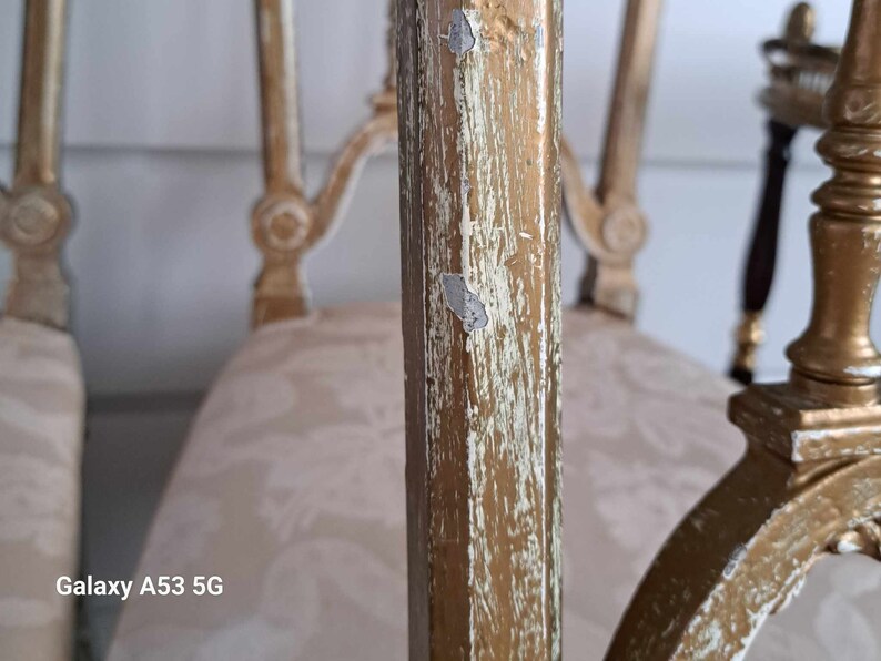 May include: Close-up of a gold-painted wooden chair with a white fabric seat. The paint is chipped and worn, revealing the wood underneath. The chair has a decorative design on the back and arms.