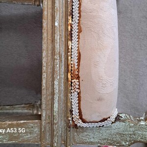 May include: Close-up of a wooden chair with a light brown fabric seat. The fabric is trimmed with a white and gold braid. The chair is distressed and has a rustic look.