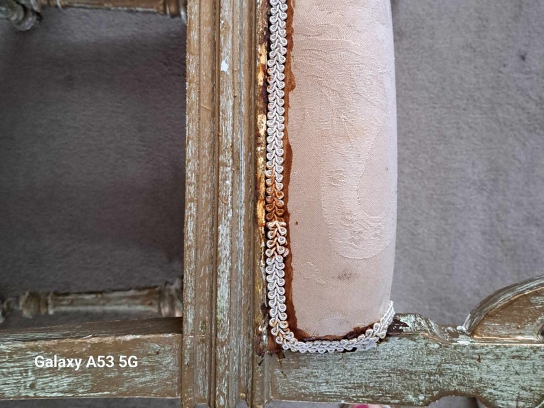 May include: Close-up of a wooden chair with a light brown fabric seat. The fabric is trimmed with a white and gold braid. The chair is distressed and has a rustic look.
