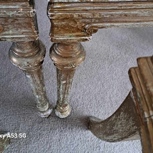 May include: Close-up of the legs of two antique chairs with distressed gold paint and a whitewash finish. The chairs have ornate carvings and a worn, vintage look.