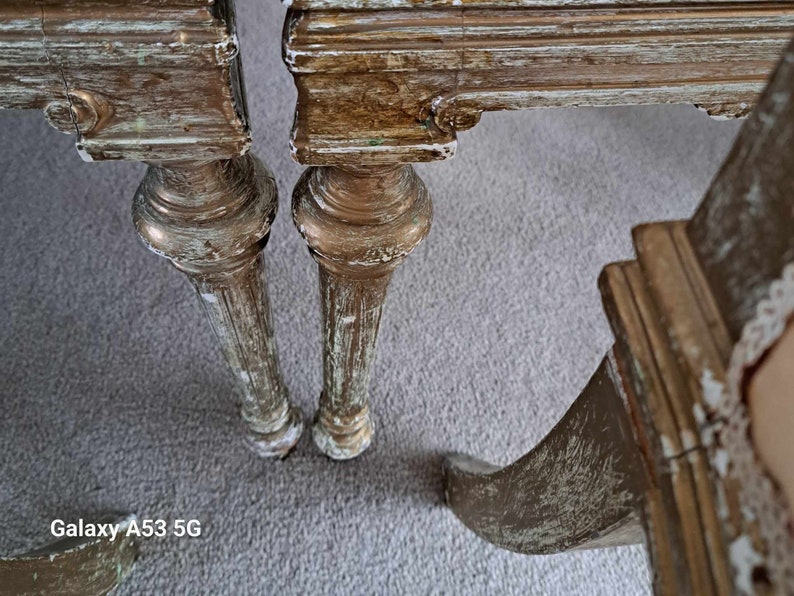May include: Close-up of the legs of two antique chairs with distressed gold paint and a whitewash finish. The chairs have ornate carvings and a worn, vintage look.