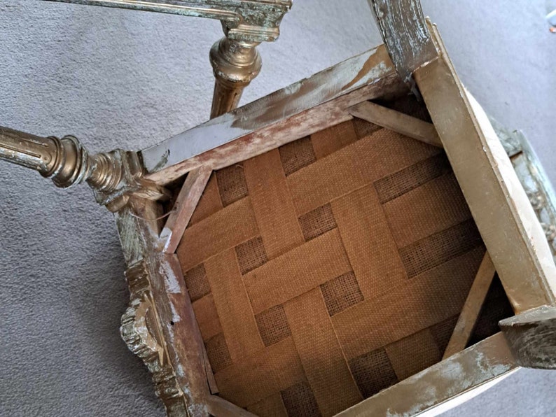 May include: The underside of a chair seat with a woven fabric seat. The chair is made of wood and has a distressed white paint finish with gold accents.