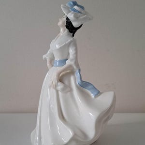 May include: A porcelain figurine of a woman wearing a white dress with a blue sash and a blue and white hat. The figurine is standing on a white surface.
