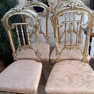 May include: Four antique chairs with ornate, carved wooden frames and gold accents. The chairs have beige fabric seats with a floral pattern.