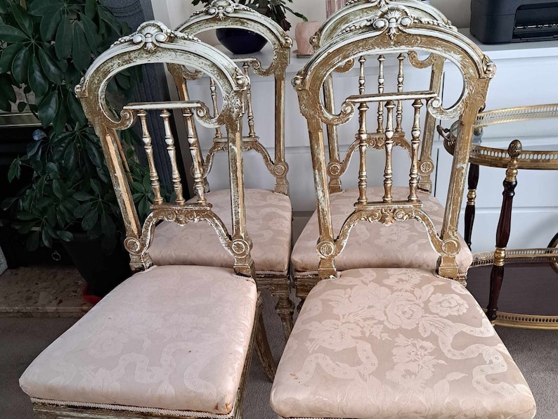 May include: Four antique chairs with ornate, carved wooden frames and gold accents. The chairs have beige fabric seats with a floral pattern.