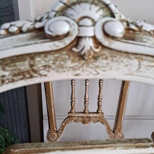 May include: A white and gold painted wooden chair with a carved back and a distressed finish. The chair has a high back with a decorative design and a curved top.