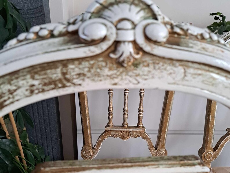May include: A white and gold painted wooden chair with a carved back and a distressed finish. The chair has a high back with a decorative design and a curved top.