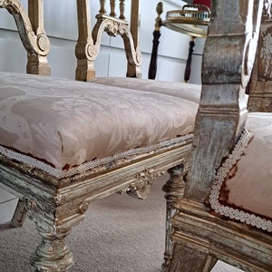 May include: Two antique chairs with ornate carved wooden frames and white painted finishes. The chairs have beige fabric seats with a delicate lace trim.
