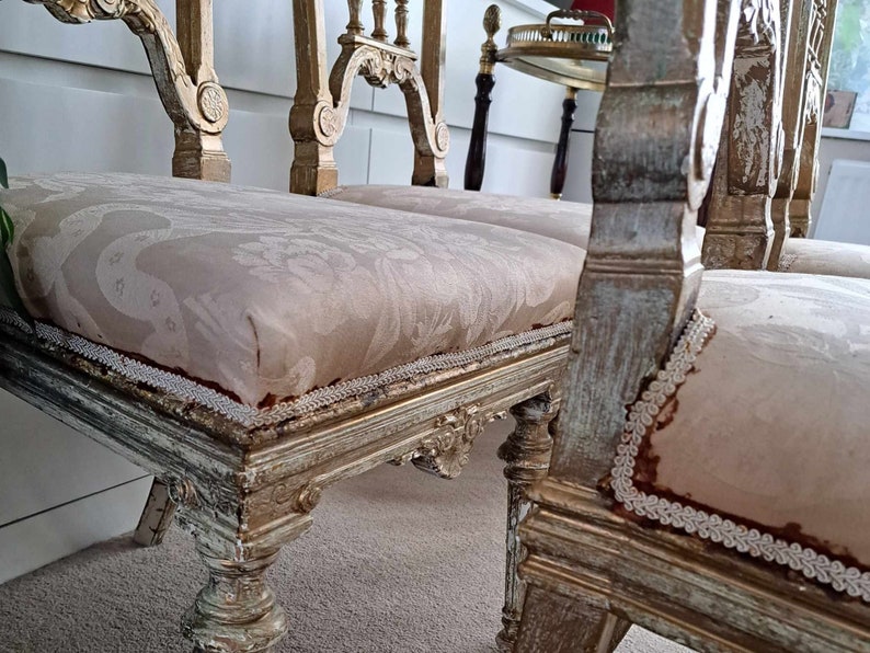 May include: Two antique chairs with ornate carved wooden frames and white painted finishes. The chairs have beige fabric seats with a delicate lace trim.