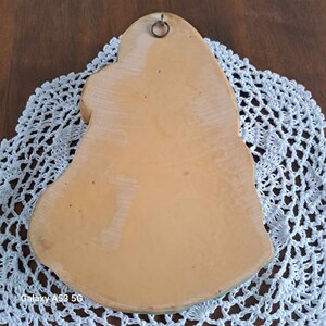 Może przedstawiać: Tył ceramicznej ozdoby ściennej w kształcie dzwonka. Dzwonek jest pomalowany w odcieniach zieleni, ż&oacute;łci i brązu. Ma metalowy haczyk do zawieszenia.