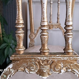 May include: A close-up of a white and gold painted chair with a carved design. The chair has a distressed finish, giving it a vintage look.