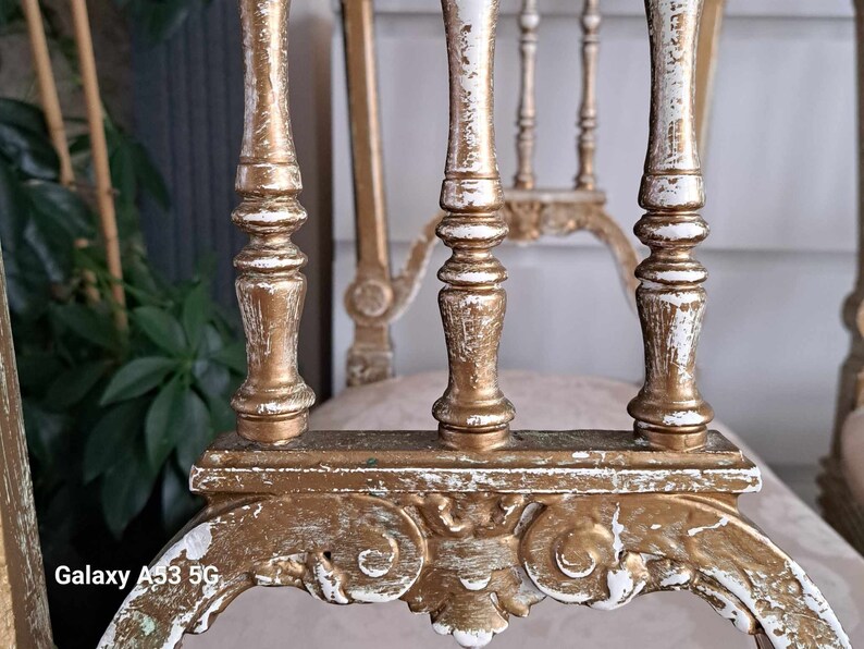 May include: A close-up of a white and gold painted chair with a carved design. The chair has a distressed finish, giving it a vintage look.
