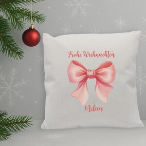 Könnte beinhalten: Weißes quadratisches Kissen mit einem rosa Schleifen-Motiv und dem Text "Frohe Weihnachten" und dem Namen "Aileen". Eine rote Weihnachtskugel hängt links oben. Der Hintergrund zeigt Schneeflockenmuster.