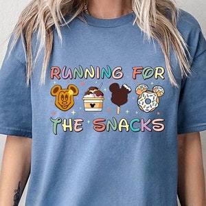 May include: A blue t-shirt with the text "Running for the Snacks" in rainbow colors. The text is surrounded by four cartoon Mickey Mouse heads, each holding a different snack: a waffle, ice cream, a Mickey Mouse-shaped ice cream bar, and a donut.